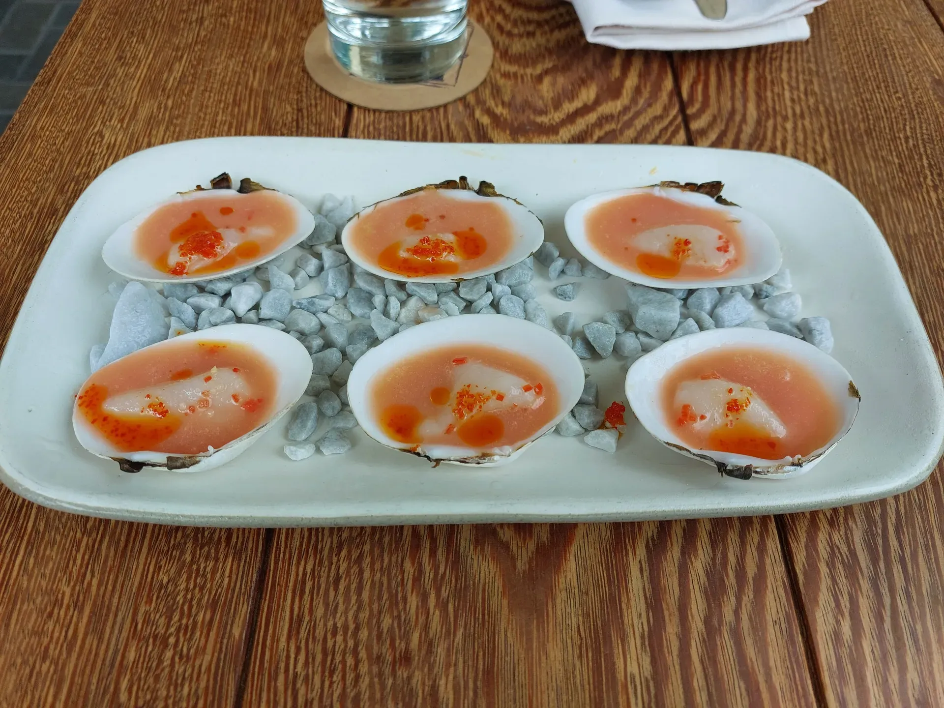 Raw seafood on clamshells over pebbles, with leche de tigre, chili oil, and roe on a white plate.