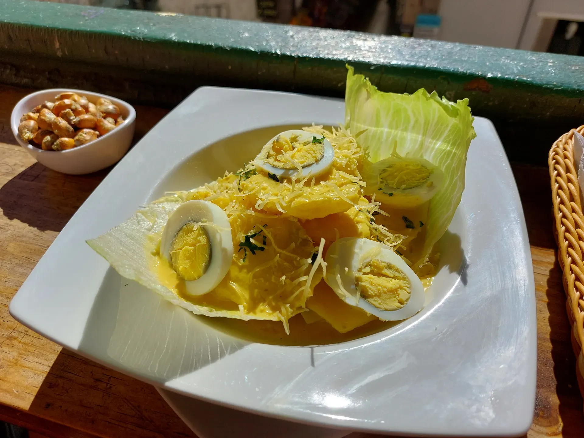 Papa a la Huancaína: sliced potatoes under creamy yellow sauce with egg, parsley, lettuce, and a bowl of toasted corn.