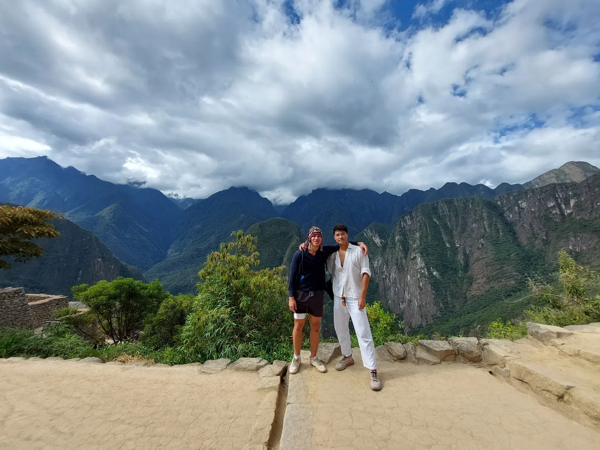 Two travelers on a stone terrace at Machu Picchu, green mountains and dramatic clouds behind them; one wears a colorful Peruvian chullo.