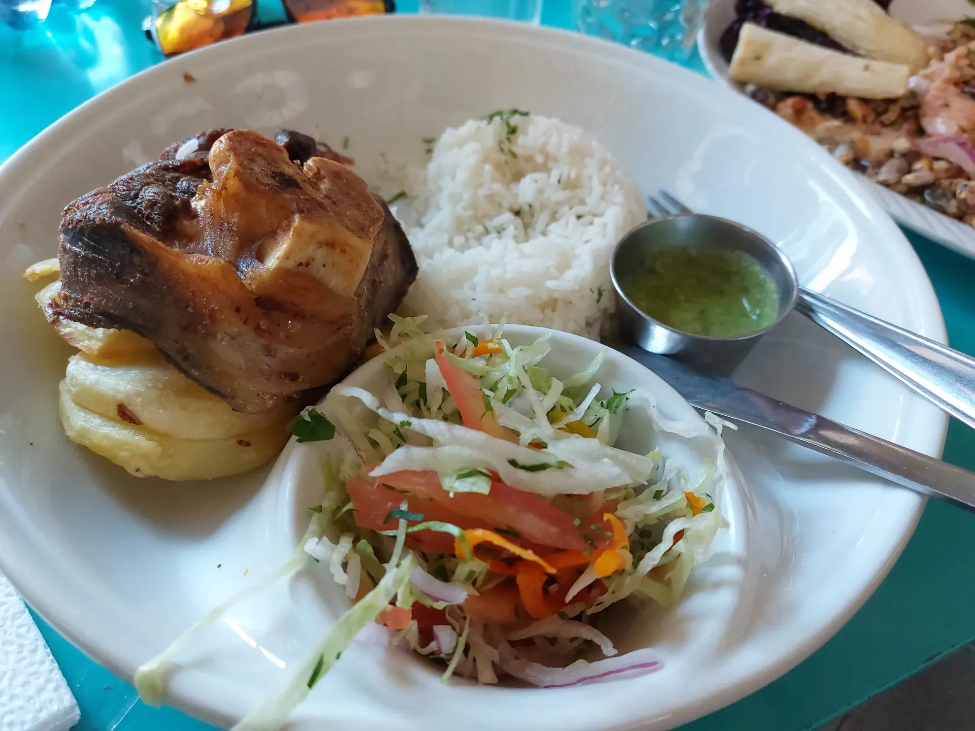 Hearty plate with roasted meat on potatoes, rice, cabbage salad, and a ramekin of green ají sauce on a turquoise table.