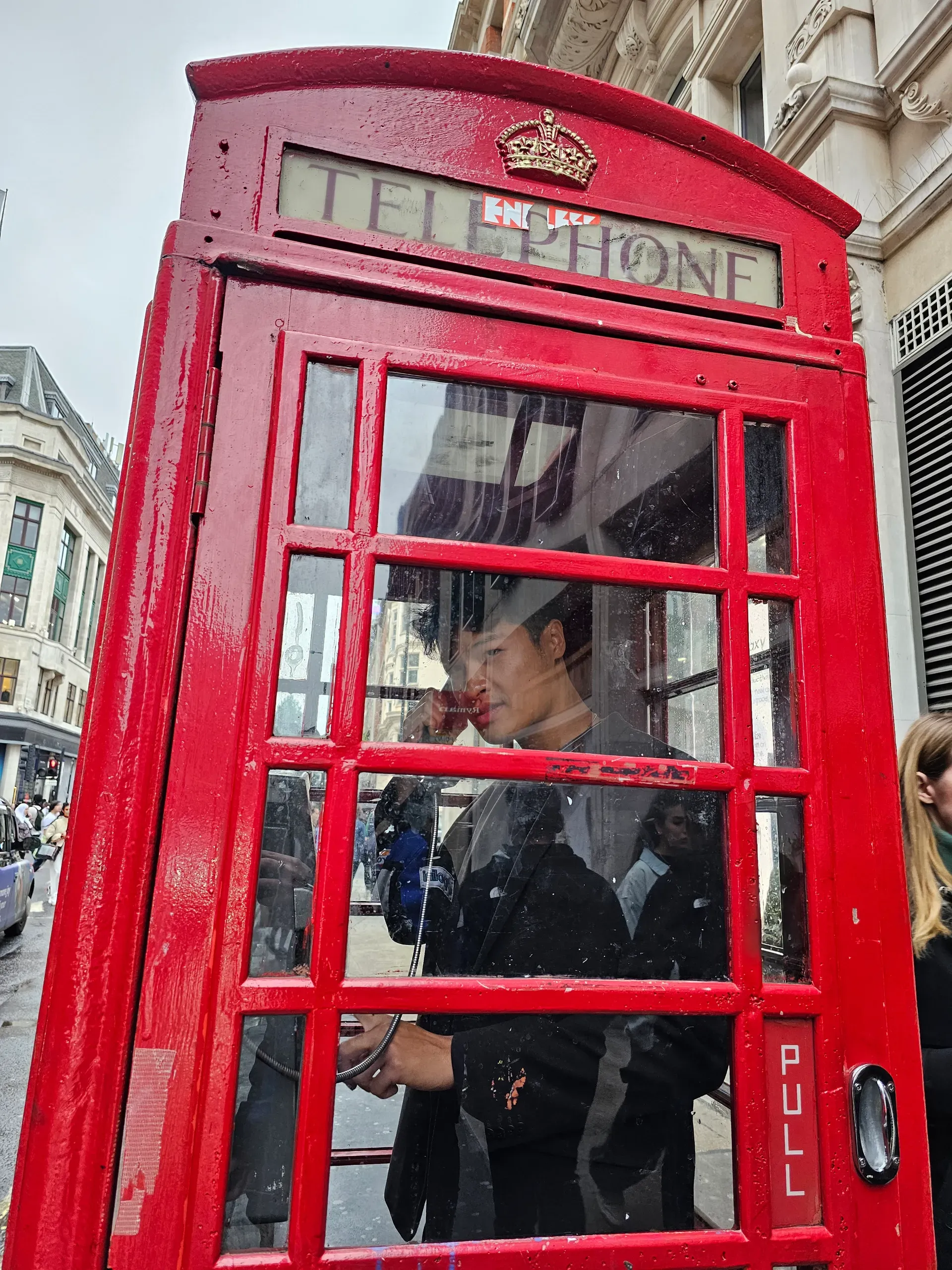 A person inside a classic red London telephone box on a grey day, holding the receiver, with stone buildings and damp pavement behind the glass.