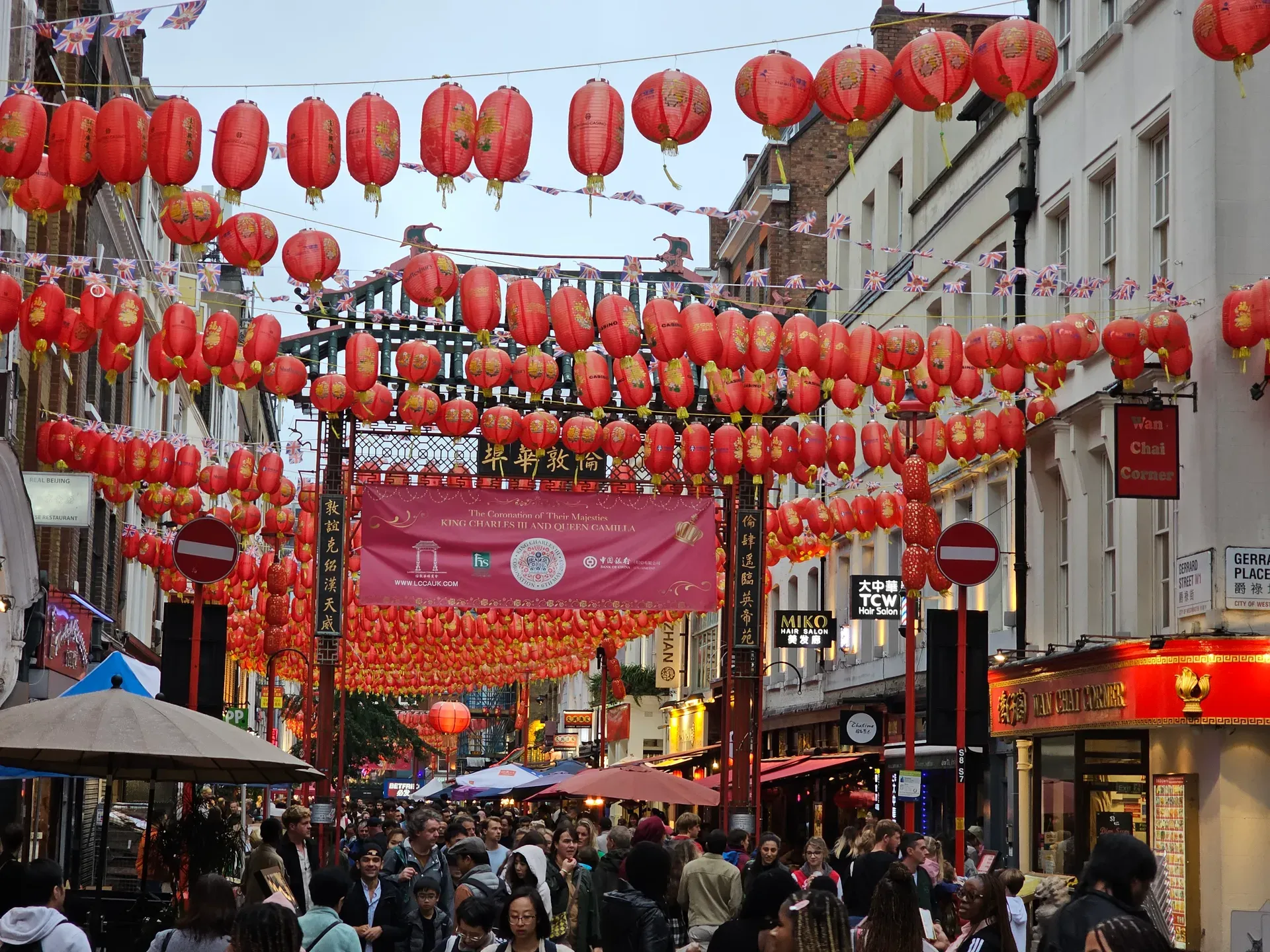 Gerrard Street in Chinatown, red lanterns overhead and a coronation banner strung across the road, crowds below under an overcast sky.