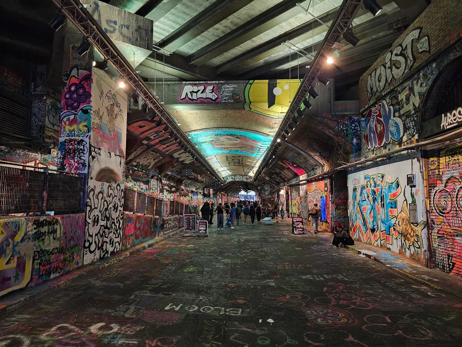 Leake Street Arches, a long graffiti tunnel under brick vaults, walls covered in layered spray paint and tags, warm lights along the ceiling.
