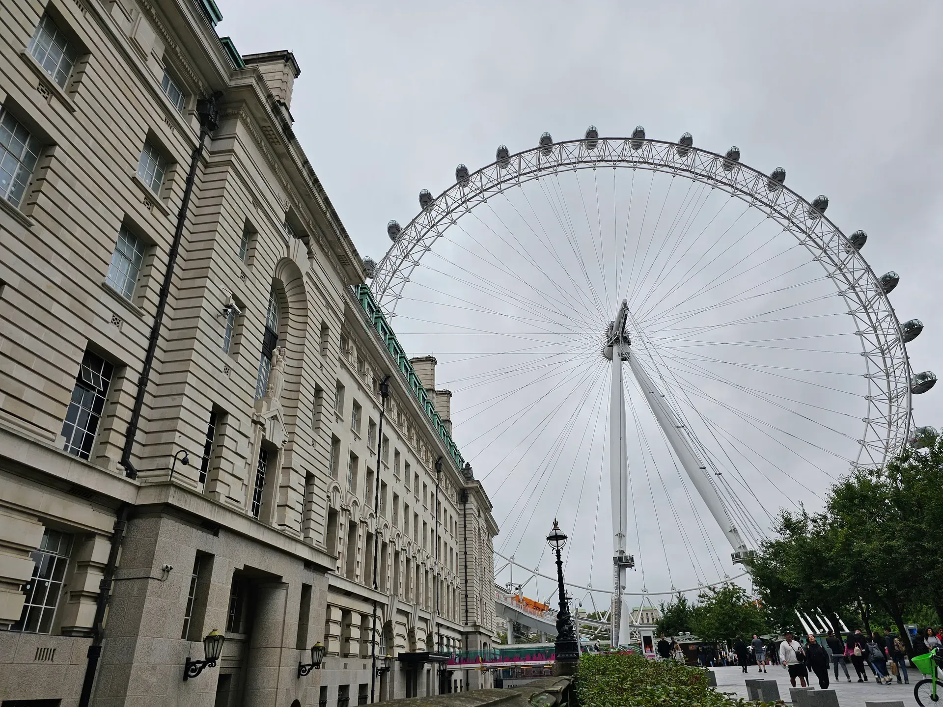 The London Eye and County Hall on an overcast day, seen from a low angle with pedestrians and cool-weather clothing in the foreground.