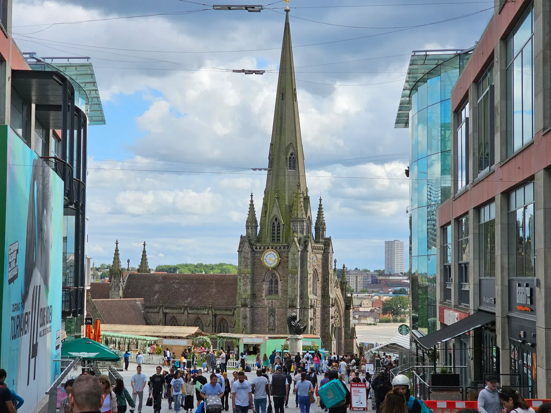 The Bull Ring on a busy afternoon: St Martin in the Bull Ring and its spire, a bronze statue, a CALM mural about suicide prevention, market stalls, and crowds between glass-fronted shops.