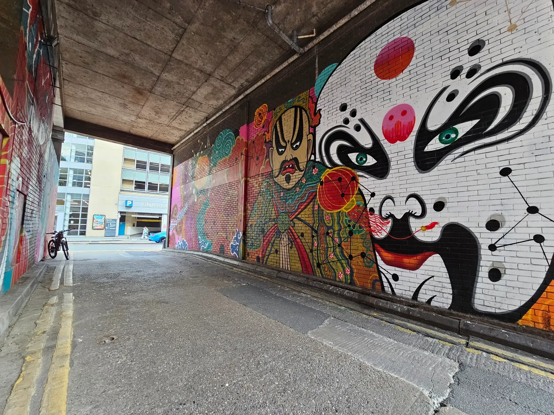 A pedestrian alley under a concrete bridge: colourful murals on brick, including a stylised face and a figure in ornate armour, with an Edgbaston car park sign visible beyond.