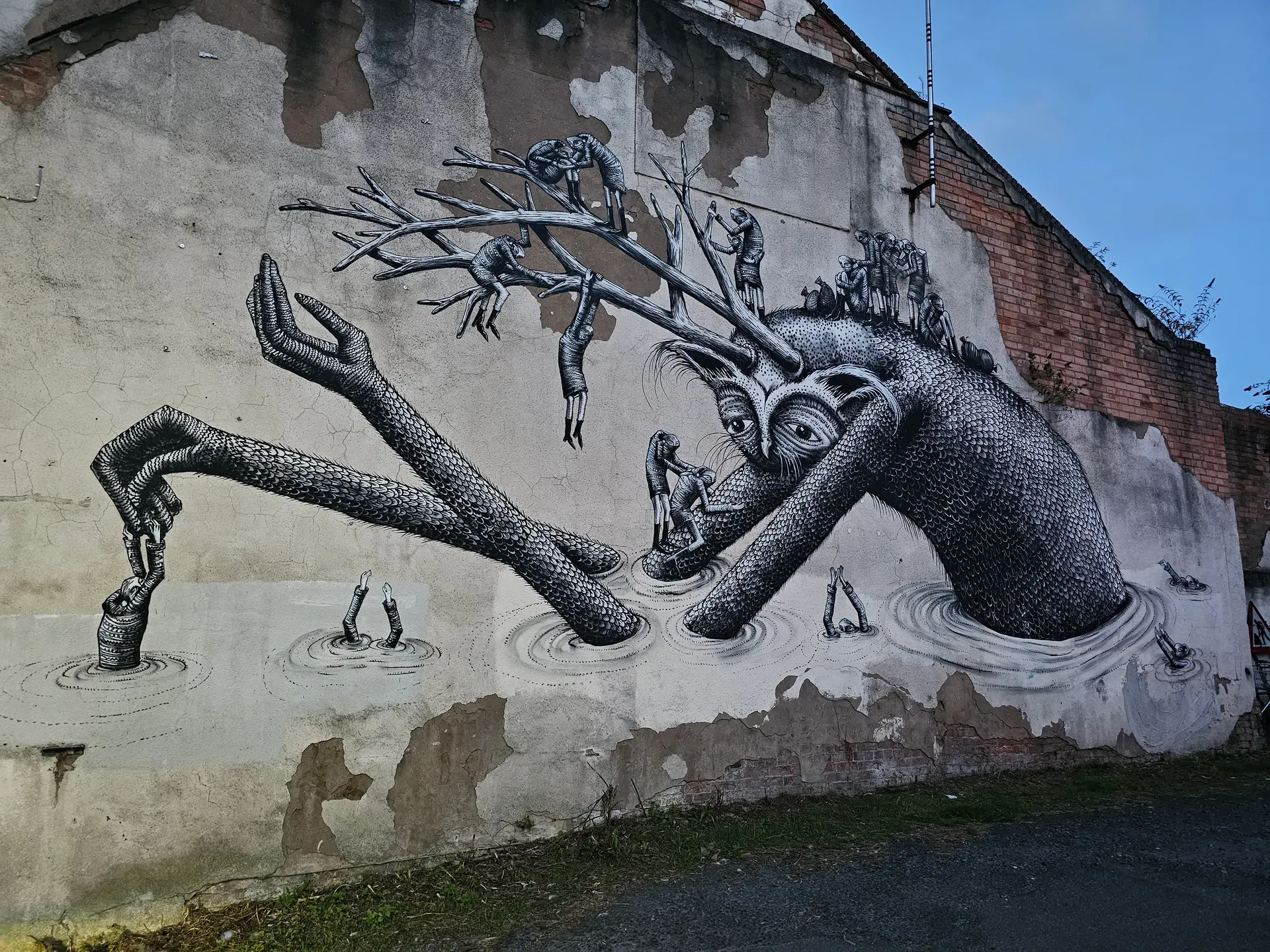 A large black-and-white mural of a fantastical owl-headed serpent with antler-like branches, small human figures, and hands emerging from drawn water, on a weathered wall with exposed brick.