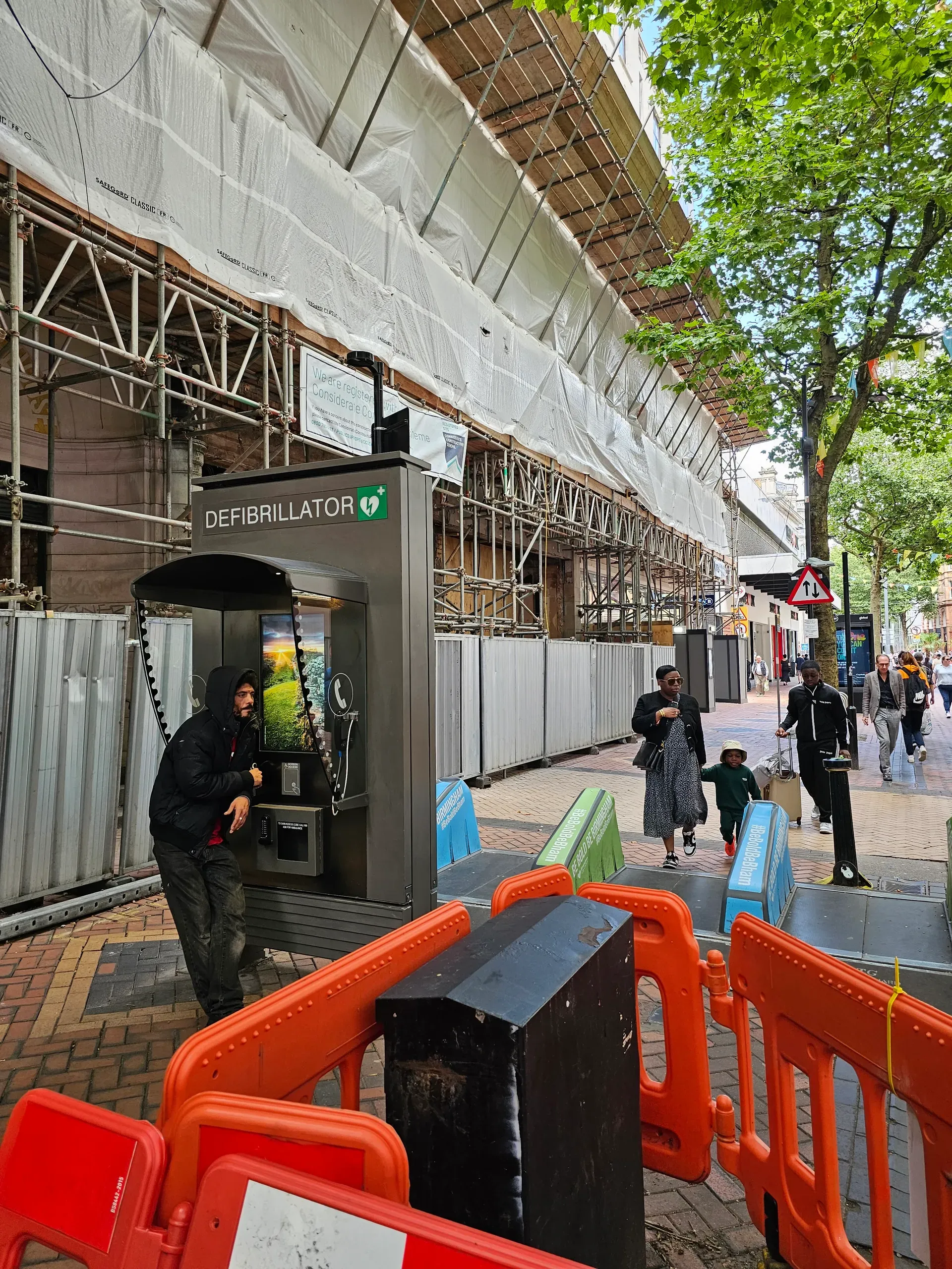 A street-side kiosk labelled DEFIBRILLATOR with a heart and lightning symbol, a curved canopy, and a screen; scaffolding and orange barriers line the pavement behind a pedestrian in a hooded jacket.