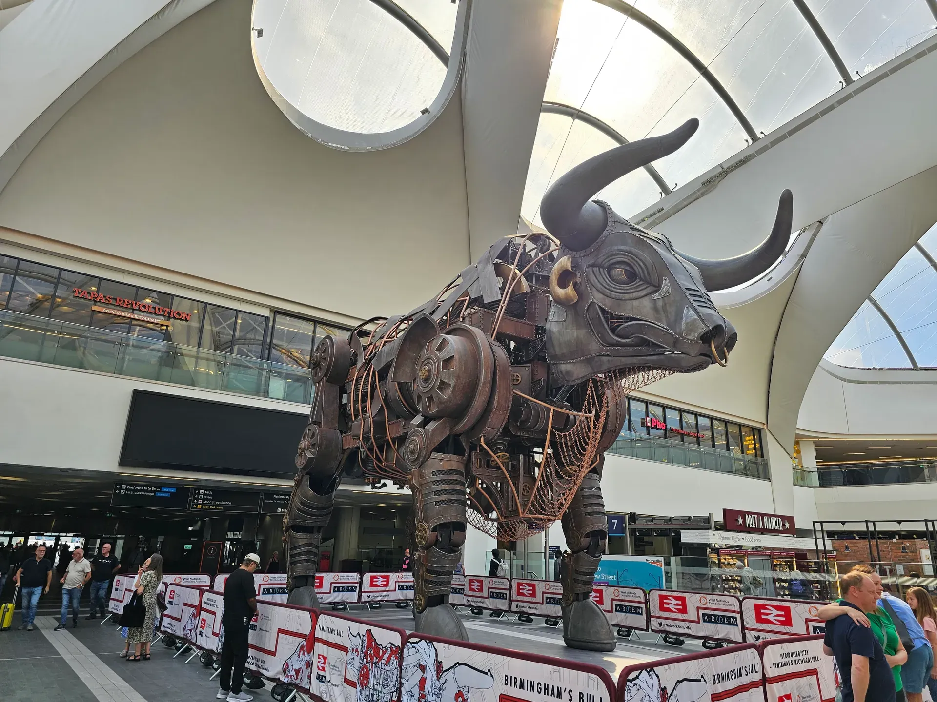 The large mechanical Raging Bull sculpture inside Birmingham New Street station, with Tapas Revolution and Pret A Manger visible and passengers crossing the concourse.