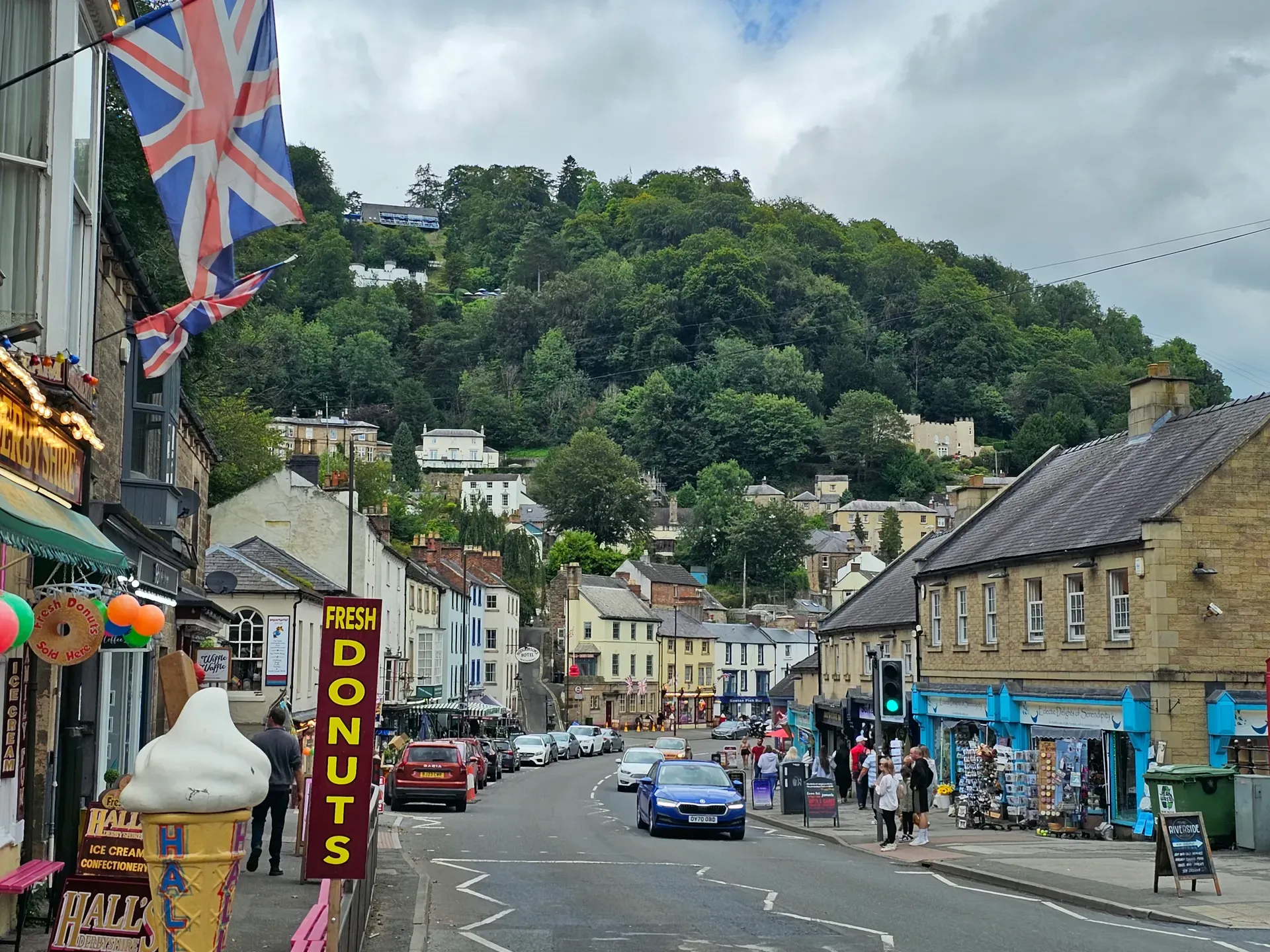 A village street with stone buildings, Union Jack flags, ice cream signs, and cars along the road.