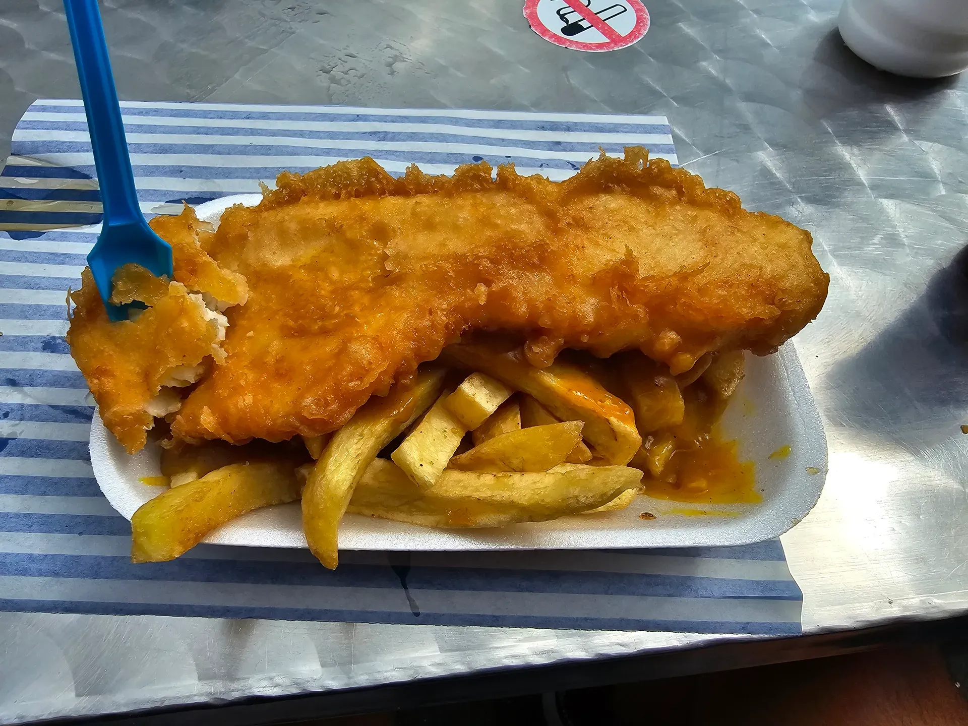A takeaway tray of battered fish and thick chips with curry sauce and a plastic fork.